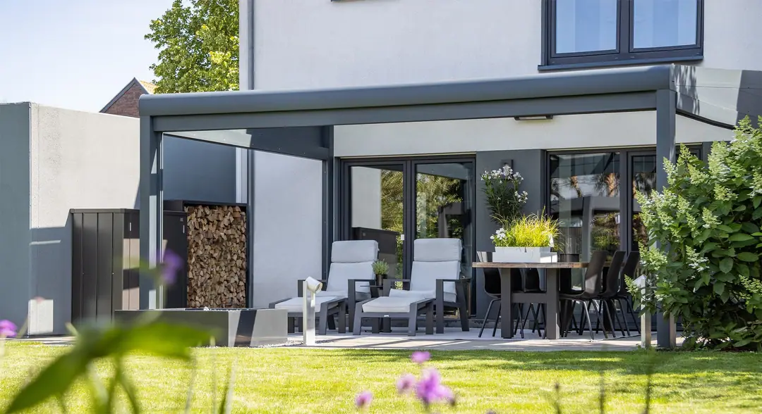 Designstyle w kolorze antracytowym przy białym domu, z boku szklany klin pod spadkiem dachu, pod dachem leżaki i stół