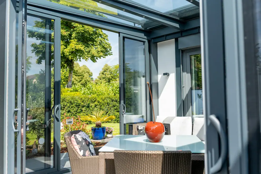 Wnętrze Ultrastyle Compact: szyby przesuwne w aluminiowej ramie z uchwytami, częściowo odsunięte na ogród, pod szklanym dachem stół i wiklinowe fotele