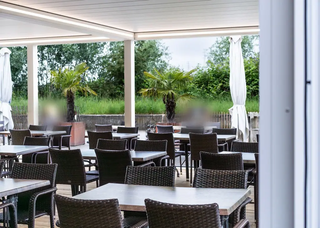 Zamknięty dach białej pergoli z taśmą LED nad ogródkiem restauracji: stoliki i wiklinowe krzesła, za słupkami palmy w donicach