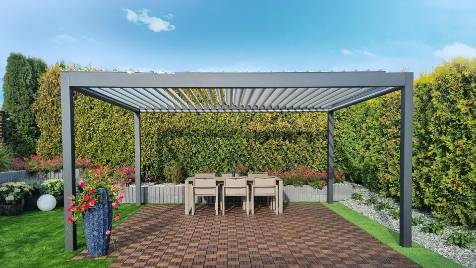 Wolnostojąca antracytowa pergola z otwartymi lamelami nad stołem z krzesłami na tarasie z płytek, wokół żywopłot i kwiaty