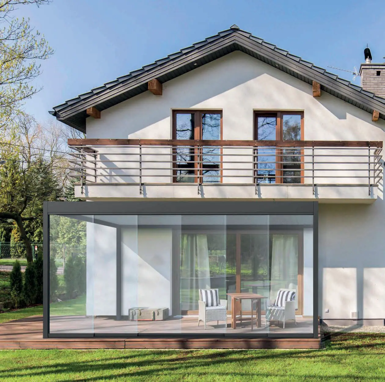 Pergola z lamelami przy domu z drewnianym balkonem, zamknięta z przodu szklanymi drzwiami przesuwnymi, w środku stolik i dwa fotele