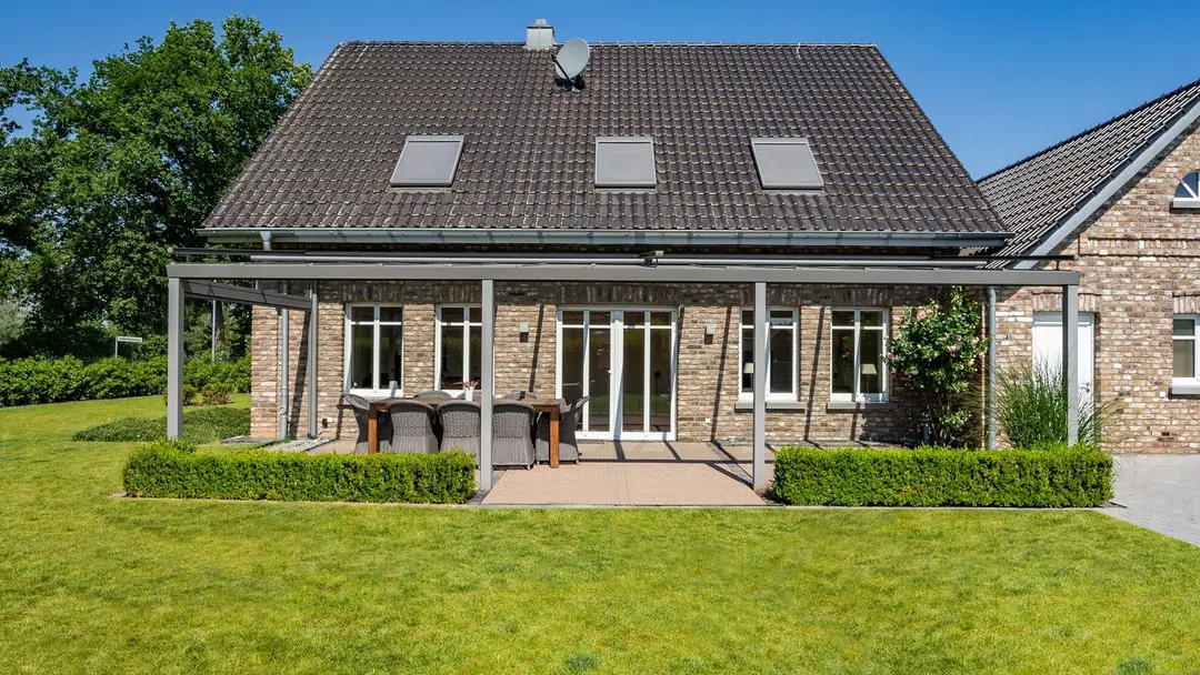 Zadaszenie Trendstyle w kolorze szarego aluminium ze szklanym dachem przy domu z cegły, przed nim żywopłot i trawnik